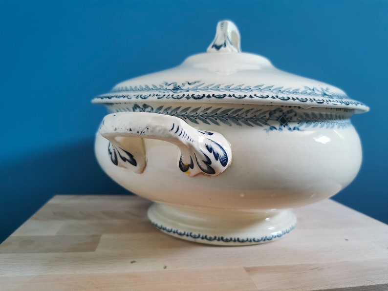 May include: A white ceramic tureen with a lid, featuring blue floral and leaf patterns. The tureen has two handles and a pedestal base. The lid has a small handle. The background is a solid blue.