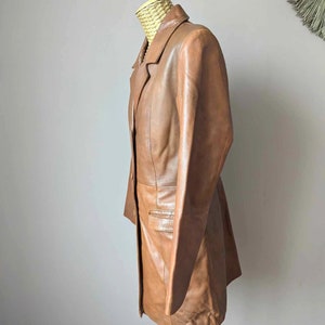 May include: A brown leather blazer displayed on a mannequin. The blazer has a classic lapel collar, a single-breasted design, and a tailored fit. The leather appears to be smooth and has a warm, medium brown tone.