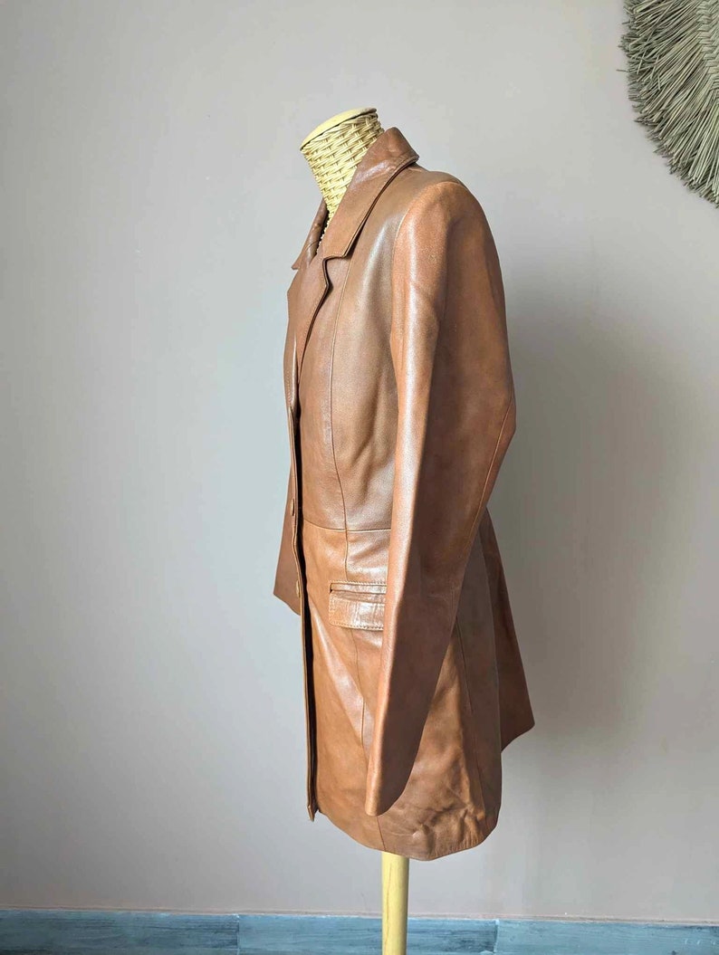 May include: A brown leather blazer displayed on a mannequin. The blazer has a classic lapel collar, a single-breasted design, and a tailored fit. The leather appears to be smooth and has a warm, medium brown tone.