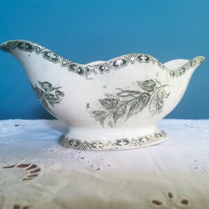 May include: A white ceramic gravy boat with a scalloped rim and pedestal base. It has a green floral and geometric pattern around the rim and base, with a floral design on the body. The gravy boat is displayed on a white lace tablecloth.