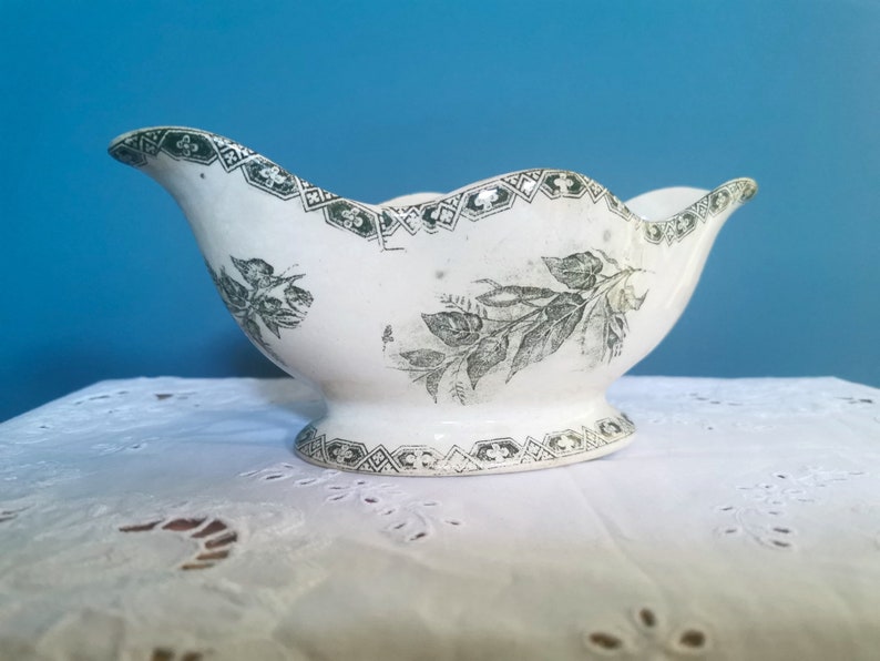 May include: A white ceramic gravy boat with a scalloped rim and pedestal base. It has a green floral and geometric pattern around the rim and base, with a floral design on the body. The gravy boat is displayed on a white lace tablecloth.