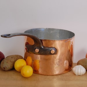 May include: A copper saucepan with a black handle and rivets. The saucepan is sitting on a wooden surface with a red onion, two yellow lemons, a garlic clove, and two potatoes.