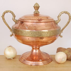 May include: A large copper serving bowl with two brass handles and a brass lid. The bowl has a decorative brass band around the top and sits on a copper pedestal.