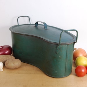 May include: A vintage green metal container with a lid and two handles. The container is shaped like a half-moon and has a slightly rusted surface. The container is sitting on a wooden surface with several vegetables, including onions, potatoes, and lemons.