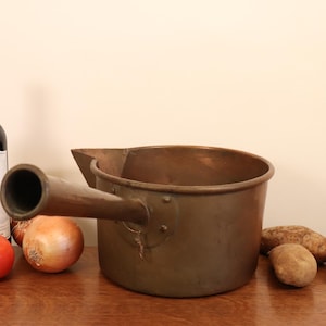 May include: A large copper pot with a long handle. The pot is sitting on a wooden table with a bottle of wine, tomatoes, and onions.