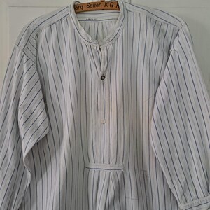 Può includere: Camicia a maniche lunghe a righe bianche e blu con colletto alla coreana e chiusura con bottoni. La camicia è appesa a un'appendiabiti di legno. La camicia ha una tasca sul davanti.