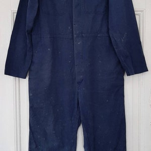 May include: A dark blue work jumpsuit with a button-up front and two large pockets on the chest. The jumpsuit has a worn and faded appearance, suggesting it has been well-used.
