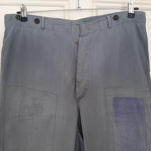 May include: A pair of gray work pants with multiple patches sewn on. The pants have a button fly and two pockets on the front.