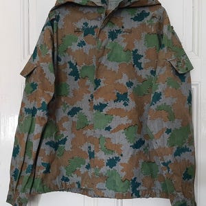 K&ouml;nnte beinhalten: Eine Kapuzenjacke in Tarnfarben mit Knopfleiste und Kordelzug in der Taille. Die Jacke ist aus leichtem Stoff und hat ein Tarnmuster in Gr&uuml;n, Braun und Grau.