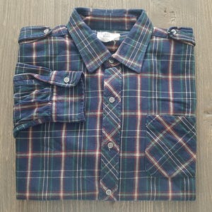 May include: A long-sleeve button-down shirt featuring a dark blue, green, and red plaid pattern. The shirt includes a pointed collar, a single chest pocket, buttoned cuffs, and shoulder epaulets with small buttons. The front placket is buttoned.