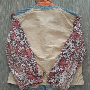 May include: A beige jacket with a floral pattern on the sleeves. The jacket has a blue denim collar and a orange collar lining. The jacket has a buckle closure at the waist.