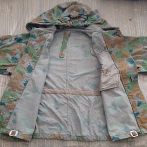 K&ouml;nnte beinhalten: Eine Tarnjacke mit Kapuze, ge&ouml;ffnet, um das Innenfutter zu zeigen. Die Jacke hat eine Innentasche mit dem aufgedruckten Text "A 2".