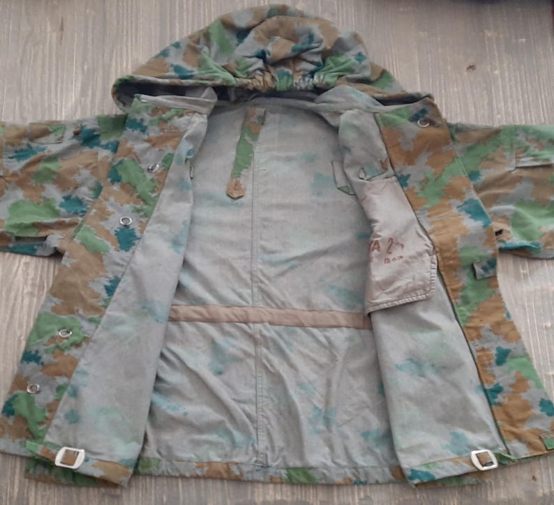 K&ouml;nnte beinhalten: Eine Tarnjacke mit Kapuze, ge&ouml;ffnet, um das Innenfutter zu zeigen. Die Jacke hat eine Innentasche mit dem aufgedruckten Text "A 2".