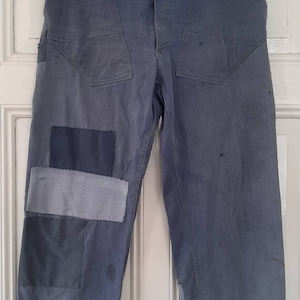 May include: A pair of blue denim work pants with multiple patches sewn on the front and back. The pants have a button fly and a dark blue waistband.