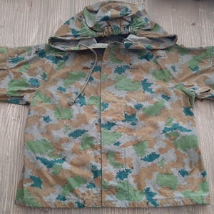 K&ouml;nnte beinhalten: Eine Kapuzenjacke in Tarnfarben mit einer Kapuze mit Kordelzug und einem Rei&szlig;verschluss. Die Jacke ist aus leichtem Stoff und hat ein Tarnmuster in Gr&uuml;n, Braun und Blau.