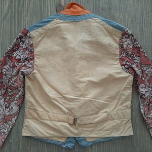 May include: A cream-colored jacket with a blue collar and cuffs. The sleeves are made of a red and white floral fabric. The jacket has a belt and a single button closure.