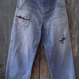 Può includere: Un paio di pantaloni da lavoro in denim blu chiaro con più tasche e segni di usura visibili. I pantaloni hanno un aspetto sbiadito e sono appesi a un appendiabiti in legno.