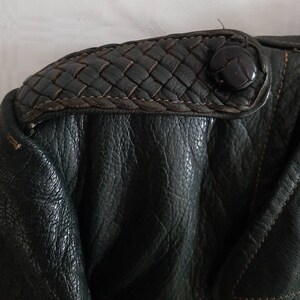 May include: Close-up of a dark green leather jacket with a woven leather detail on the shoulder. The detail has a black button.
