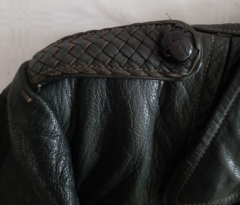 May include: Close-up of a dark green leather jacket with a woven leather detail on the shoulder. The detail has a black button.