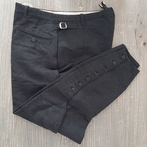Puede incluir: Un pantalón de montar de color gris oscuro con cierre de botón en el lateral. El pantalón tiene una trabilla para el cinturón y un bolsillo trasero.