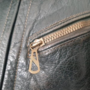 May include: Close-up of a zipper on a dark green leather jacket. The zipper pull is a metal loop with a decorative design.