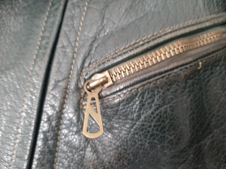 May include: Close-up of a zipper on a dark green leather jacket. The zipper pull is a metal loop with a decorative design.