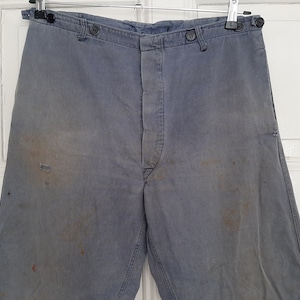 Könnte beinhalten: Verblasste blaue Arbeitshose mit Knopfverschlüssen und hoher Taille. Die Hose weist Gebrauchsspuren mit Flecken und einem kleinen Riss auf. Die Hose hängt an einem Metallbügel vor weißem Hintergrund.
