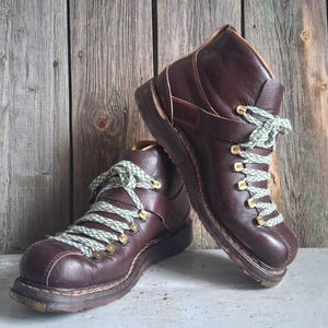 May include: A pair of dark brown leather ankle boots featuring a textured finish, gold-toned metal eyelets, and light green and white patterned laces. The boots have a rounded toe, a thick dark sole, and a strap detail across the mid-foot area.