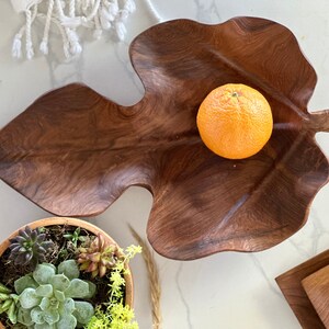 May include: A wooden leaf-shaped bowl with a smooth, dark brown finish. An orange sits in the centre of the bowl.