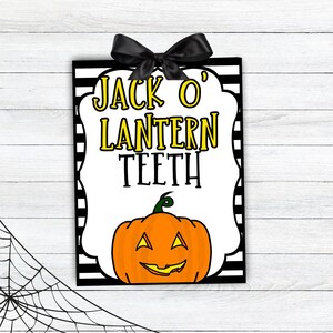 Peut inclure: Une pancarte à rayures noires et blanches avec une bordure blanche et un nœud noir. La pancarte indique "Jack O' Lantern Teeth" en lettres jaunes et présente une citrouille orange de dessin animé avec un sourire aux dents.
