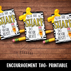 Puede incluir: Tres etiquetas de ánimo con lazos amarillos y lápices. Cada etiqueta tiene el texto "You are so SHARP, DO YOUR BEST!" y un personaje de lápiz de dibujos animados. Las etiquetas están sobre un fondo de madera.