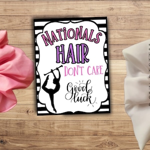 May include: A black and white striped sign with the text "Nationals Hair Don't Care" in pink and purple. A silhouette of a figure holding a baton is in the center of the sign. The text "Good Luck" is written in cursive below the figure.