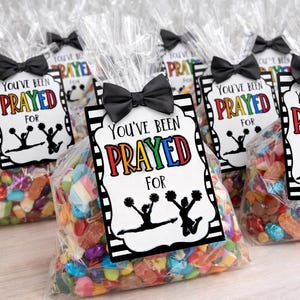 May include: Clear cellophane bags filled with colorful candies, each topped with a black bow and a tag that reads "You've Been Prayed For." The tags feature a black and white design with cheerleaders.