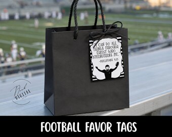 May include: A matte black paper gift bag with black twisted rope handles. Attached to the handle is a rectangular white tag with black stripes, featuring a silhouette of a football player and the text I CAN DO ALL THINGS THROUGH CHRIST WHO STRENGTHENS ME. -PHILLIPIANS 4:13.