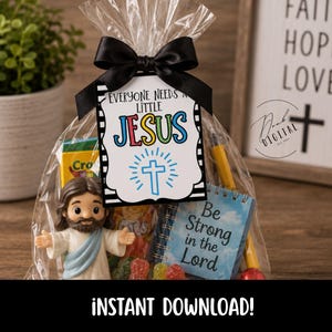 May include: A clear plastic gift bag tied with a black ribbon, containing a small Jesus figurine, a box of crayons, a blue notebook with the text Be Strong in the Lord, and colorful candies. A tag reads EVERYONE NEEDS A LITTLE JESUS with a cross.