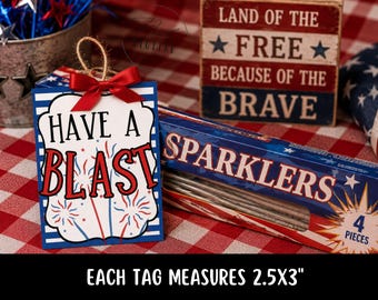 May include: A patriotic-themed gift tag with the words "HAVE A BLAST" in red and blue, adorned with a red bow and a twine loop. The tag measures 2.5 x 3 inches. A box of sparklers and a wooden sign that reads "LAND OF THE FREE BECAUSE OF THE BRAVE" are also visible.