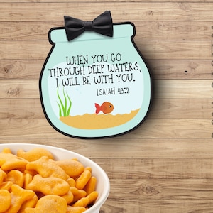 May include: A blue and white fishbowl with a gold fish inside. The fishbowl has a black bow tie on top and the text "WHEN YOU GO THROUGH DEEP WATERS, I WILL BE WITH YOU. ISAIAH 43:2" printed on it.
