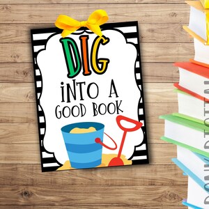 Op de afbeelding: Een kleurrijk bord met de tekst "DIG INTO A GOOD BOOK" in speelse letters. Het bord toont een illustratie van een strandemmer en schop, met een geel lint aan de bovenkant. Een stapel boeken is zichtbaar op de achtergrond.