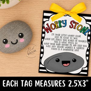 May include: A gray pebble with a painted smiling face and pink cheeks next to a rectangular tag featuring black and white stripes, a yellow ribbon, and the text: WORRY STONE, I'M YOUR LITTLE WORRY STONE, KEEP ME CLOSE TO YOU. Tag measures 2.5x3 inches.