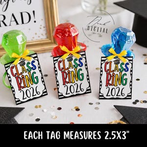 May include: Three graduation ring favors in green, red, and blue, each with a yellow ribbon and a tag that reads "Class Ring 2026." The tags measure 2.5x3 inches. A graduation cap and a framed sign that says "Congrats Grad!" are in the background.