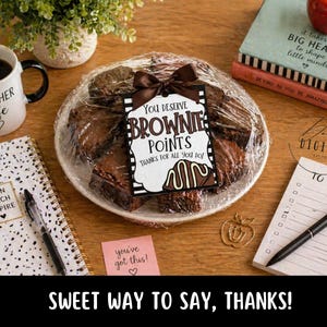 May include: A plate of brownies wrapped in plastic, tied with a brown ribbon, and a tag that reads "You Deserve Brownie Points." A mug with "Teacher Life" and a notebook are on a wooden surface. A red apple and books are also present.