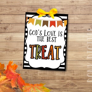 May include: A rectangular sign with the text "GOD'S LOVE IS THE BEST TREAT" in black and orange lettering. The sign has a white border with black stripes and a yellow bow at the top. Colourful autumn leaves are in the lower left corner.