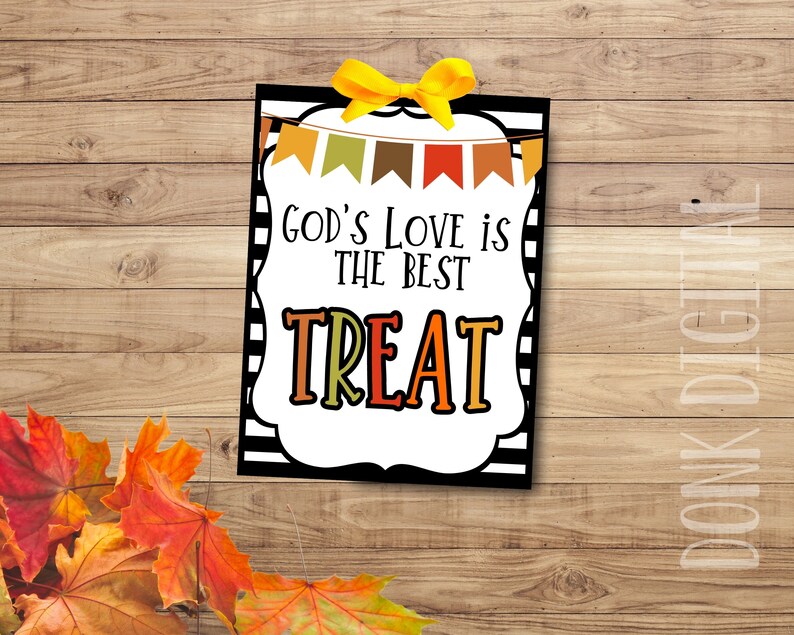 May include: A rectangular sign with the text "GOD'S LOVE IS THE BEST TREAT" in black and orange lettering. The sign has a white border with black stripes and a yellow bow at the top. Colourful autumn leaves are in the lower left corner.