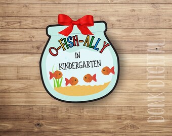 Puede incluir: Un colgador de puerta en forma de pecera azul y blanca con un lazo rojo. La pecera tiene el texto "O-FISH-ALLY IN KINDERGARTEN" y cuatro peces de colores naranja dentro.