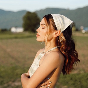 May include: A woman with long, wavy brown hair is wearing a white and green plaid dress and a white and green plaid bandana. She is standing in a field with green grass and mountains in the background.