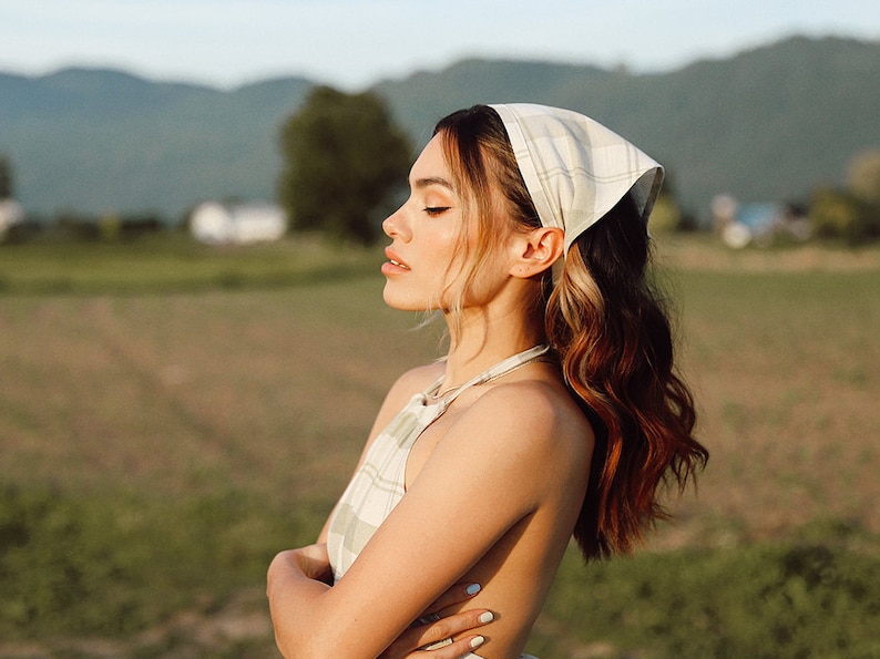 May include: A woman with long, wavy brown hair is wearing a white and green plaid dress and a white and green plaid bandana. She is standing in a field with green grass and mountains in the background.