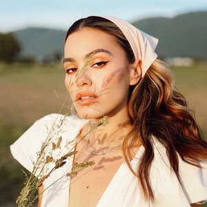 May include: A woman with long brown hair wearing a white dress and a pink bandana. She is holding a bouquet of dried flowers and looking at the camera.