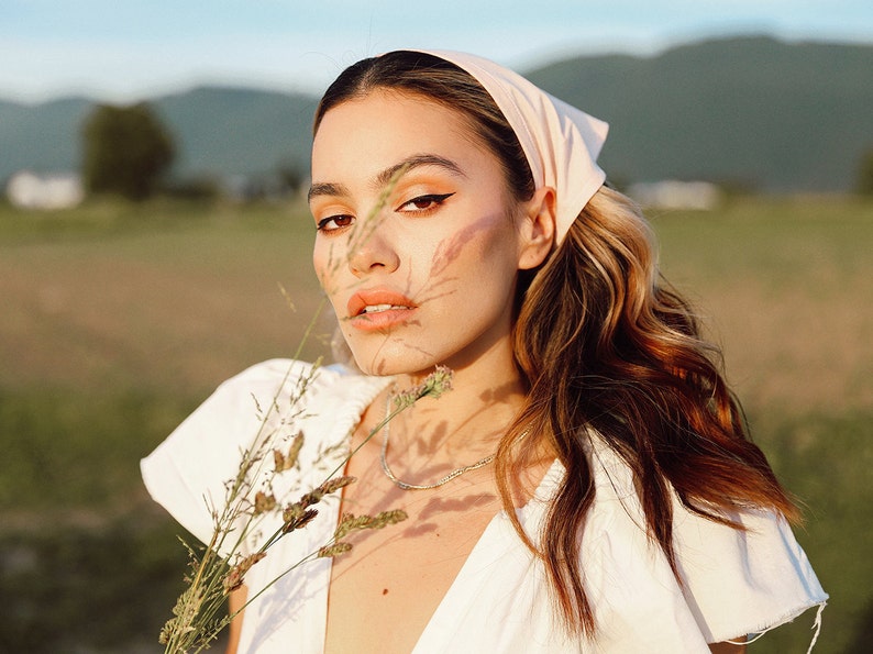 May include: A woman with long brown hair wearing a white dress and a pink bandana. She is holding a bouquet of dried flowers and looking at the camera.