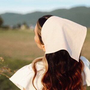 May include: A woman with long brown hair wearing a white linen headscarf and a white dress, standing in a field.