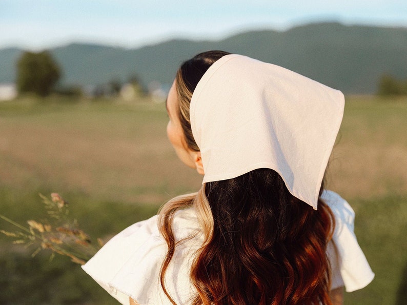 May include: A woman with long brown hair wearing a white linen headscarf and a white dress, standing in a field.